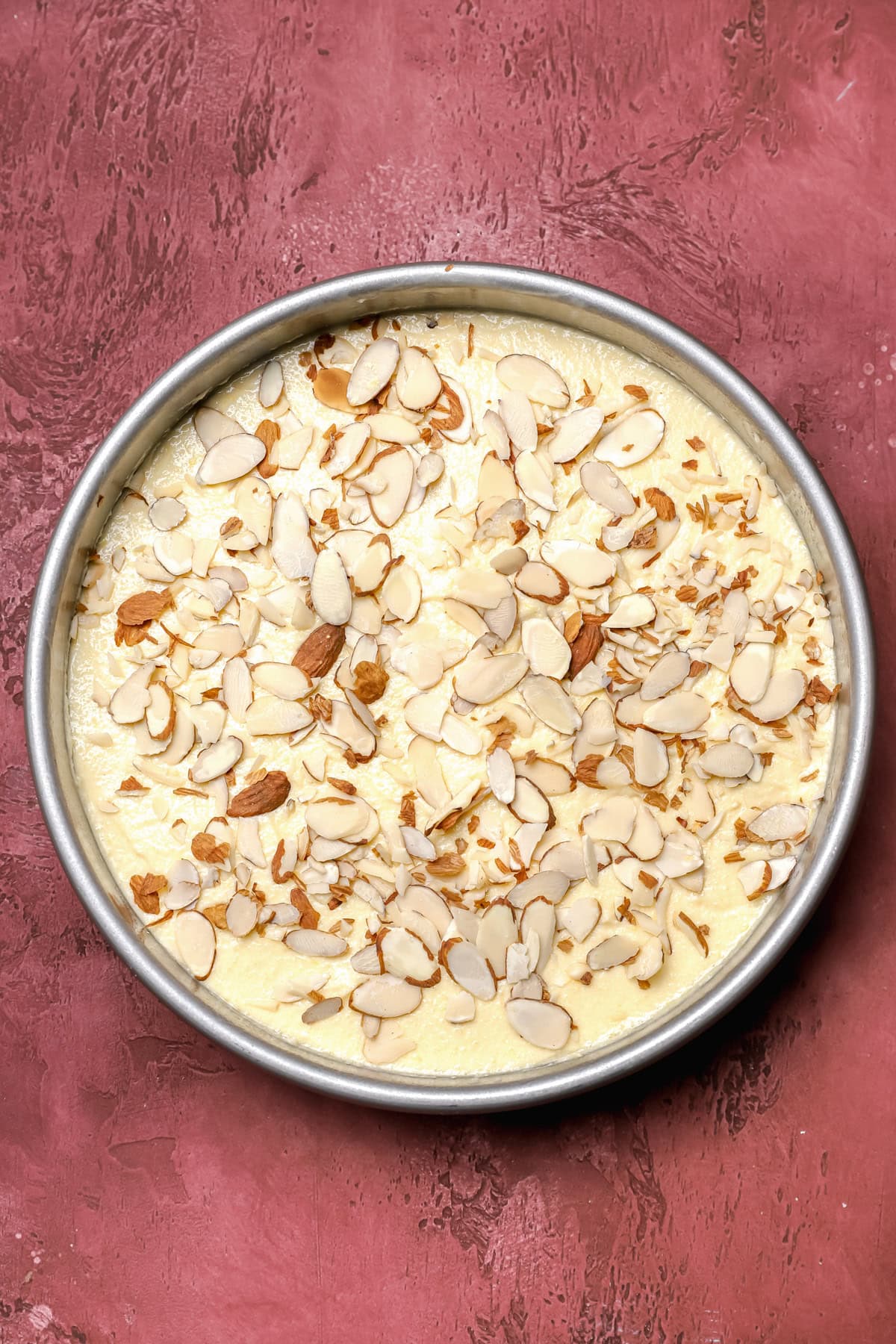 A round cake pan filled with unbaked cake batter topped with sliced almonds, placed on a textured, dark pink surface.