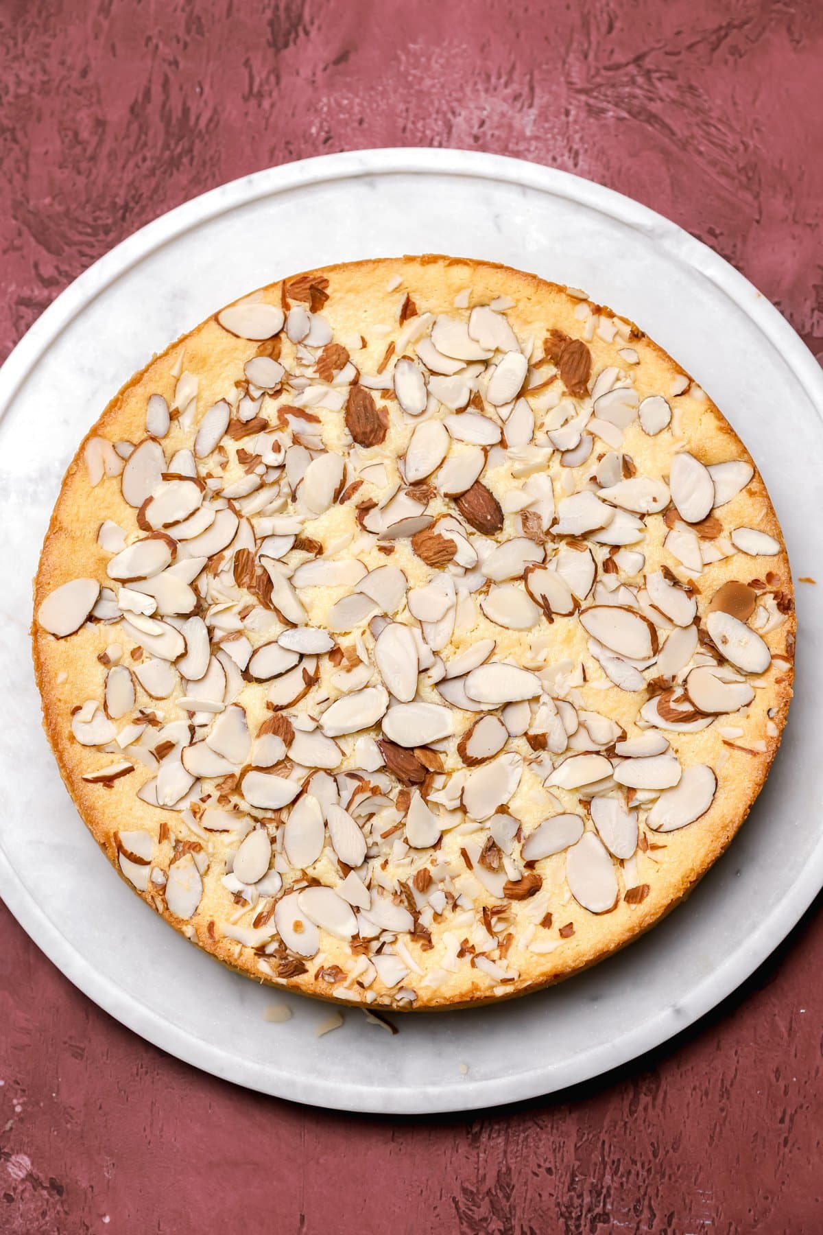 A round almond cake topped with sliced almonds, displayed on a white plate over a textured reddish-brown surface.