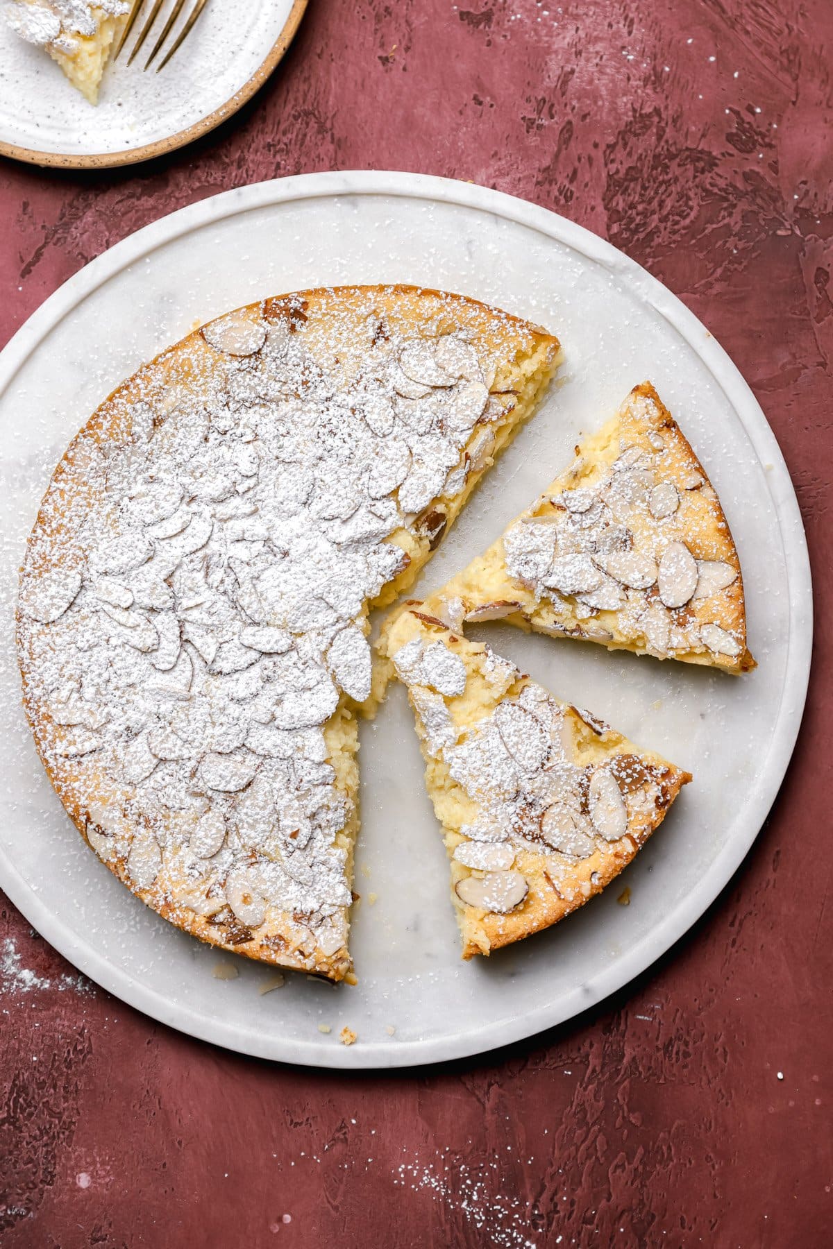 A round almond cake dusted with powdered sugar and topped with sliced almonds sits on a white plate. Two slices have been cut out, and a fork rests on a small plate in the corner. The background is a textured red surface.