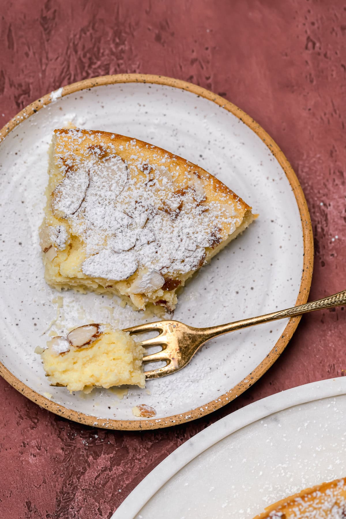 A slice of almond cake topped with powdered sugar sits on a speckled plate. A gold fork holds a small bite of cake, and the background is a textured reddish surface.