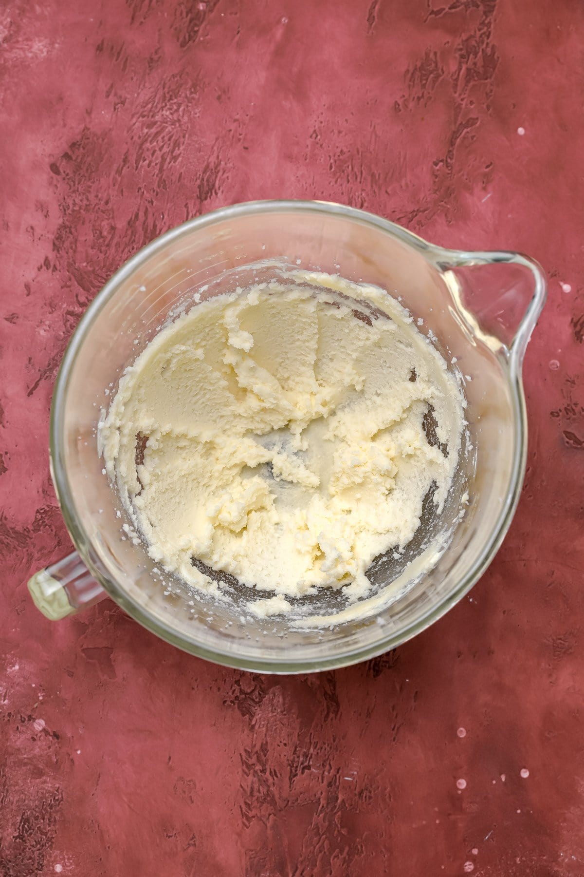 A glass mixing bowl filled with creamed butter and sugar sits on a textured reddish-pink surface.