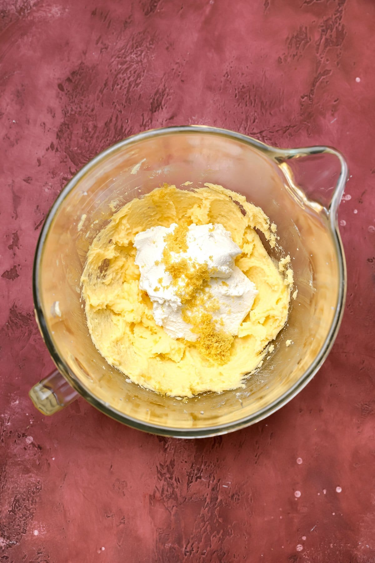 A glass mixing bowl on a maroon surface, containing a creamy yellow mixture topped with white cream and a sprinkling of lemon zest.