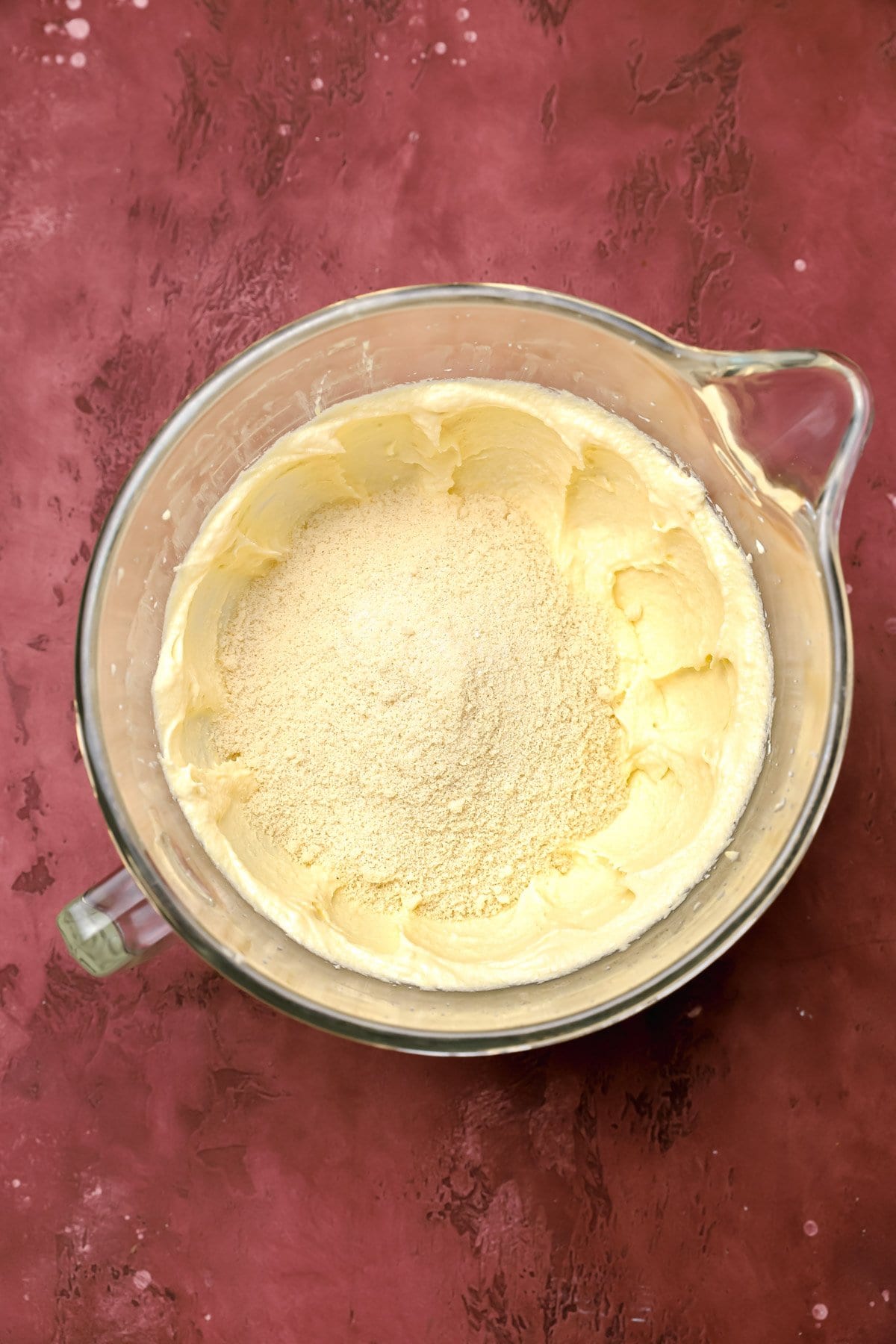 A glass mixing bowl on a maroon surface, containing pale yellow creamed butter and sugar with a mound of flour sitting on top, ready to be mixed together.