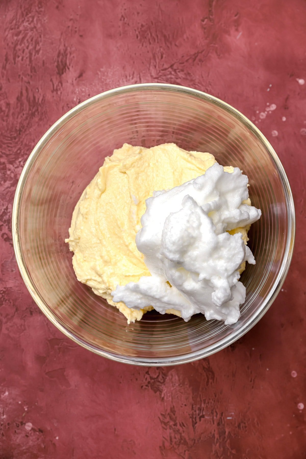 A glass bowl on a maroon surface contains a yellow batter and a dollop of fluffy whipped egg whites.
