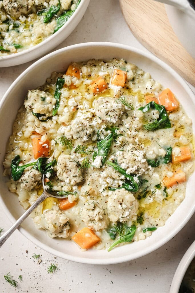 A bowl of creamy soup with spinach, diced carrots, rice, and herb-seasoned meatballs, garnished with fresh dill. A spoon rests in the bowl, and part of another bowl is visible in the background.