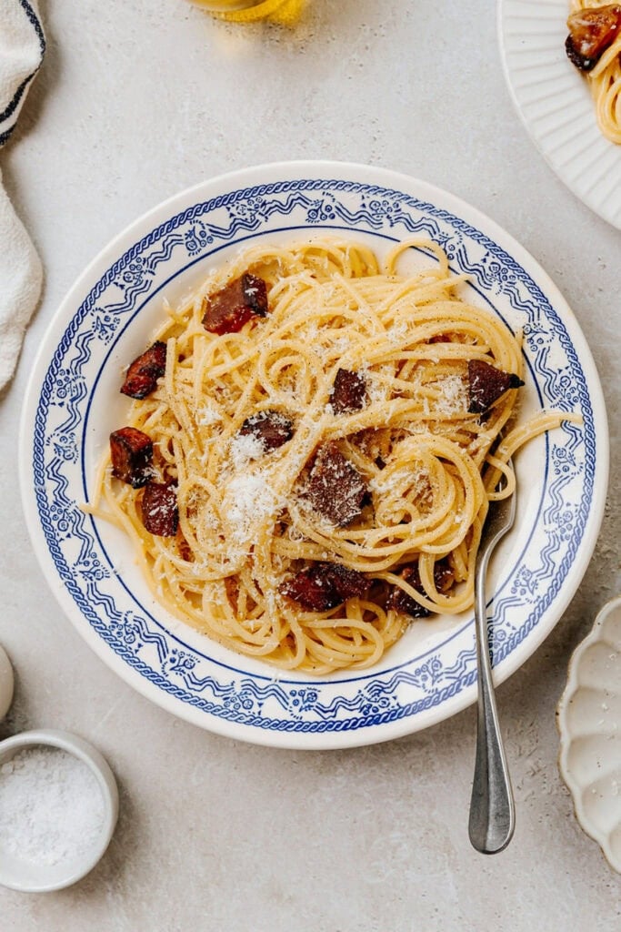 A bowl of spaghetti carbonara topped with grated cheese and crispy pancetta pieces, served in a white and blue patterned dish with a fork resting inside.
