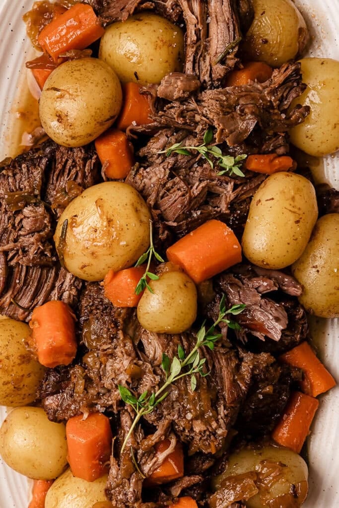 A close-up of pot roast with tender beef, whole baby potatoes, and chopped carrots, garnished with fresh thyme on a white serving platter.
