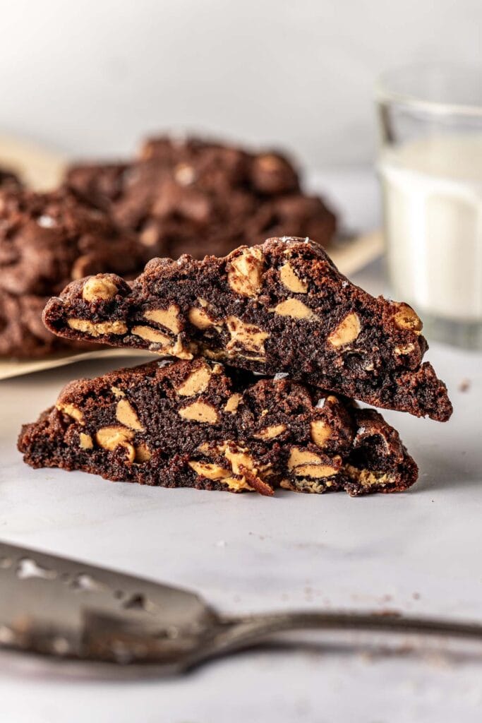 A close-up of two thick, richly chocolate cookies with visible peanut butter chips, stacked on a marble surface. Blurred cookies and a glass of milk are in the background.