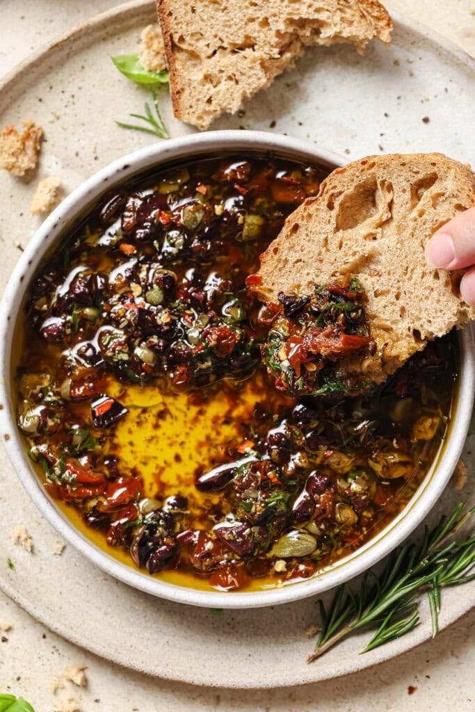 A hand dips a piece of rustic bread into a bowl of olive oil mixed with herbs and sun-dried tomatoes. The bowl sits on a light plate, garnished with fresh rosemary and basil.