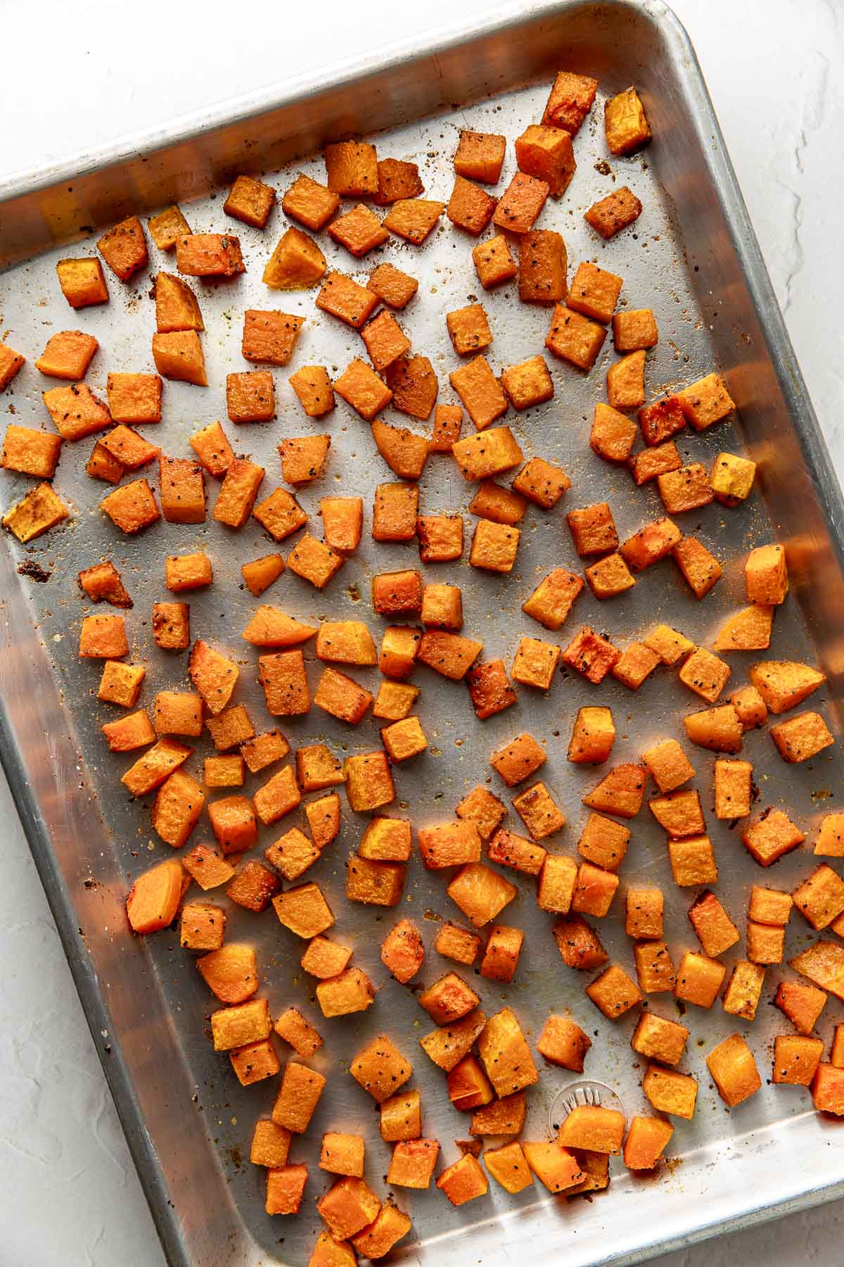 Cubed pieces of roasted sweet potato, evenly spread out on a metal baking sheet. The sweet potatoes are golden brown with a light seasoning. The baking sheet rests on a white surface.
