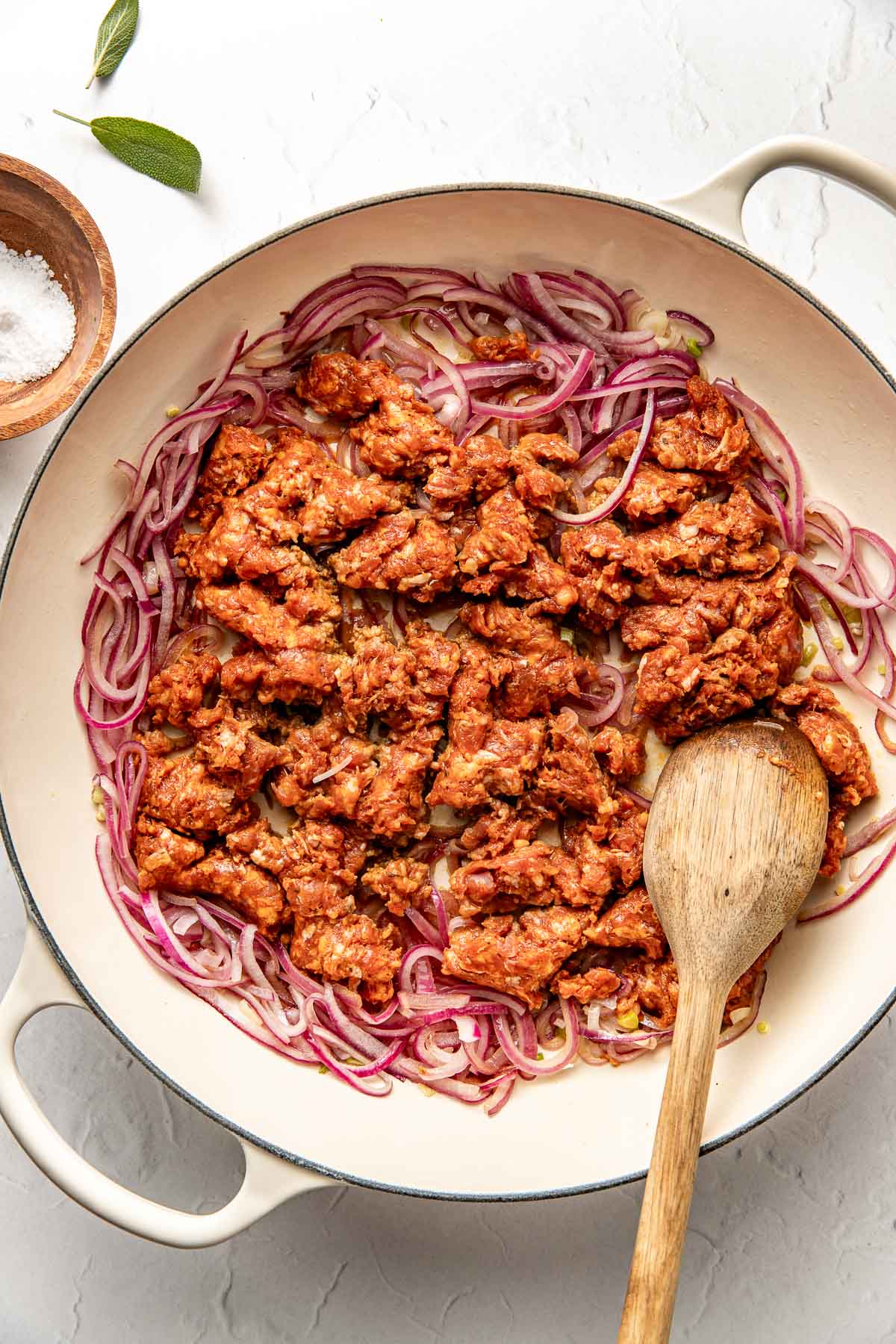 A large white skillet contains sliced red onions and crumbled sausage, being sautéed together. A wooden spoon rests in the pan, and a small bowl of salt sits nearby on the white countertop.