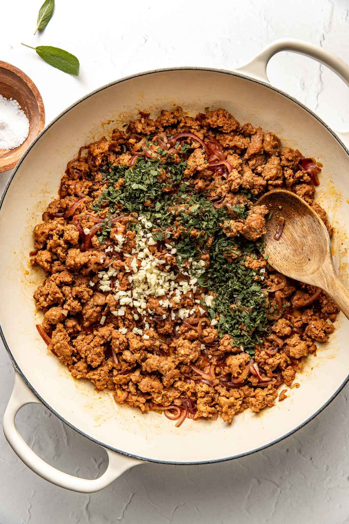 A white skillet filled with cooked ground meat, chopped herbs, and minced garlic, with a wooden spoon resting on the side. A small bowl of salt and sage leaves are nearby on a light-colored surface.