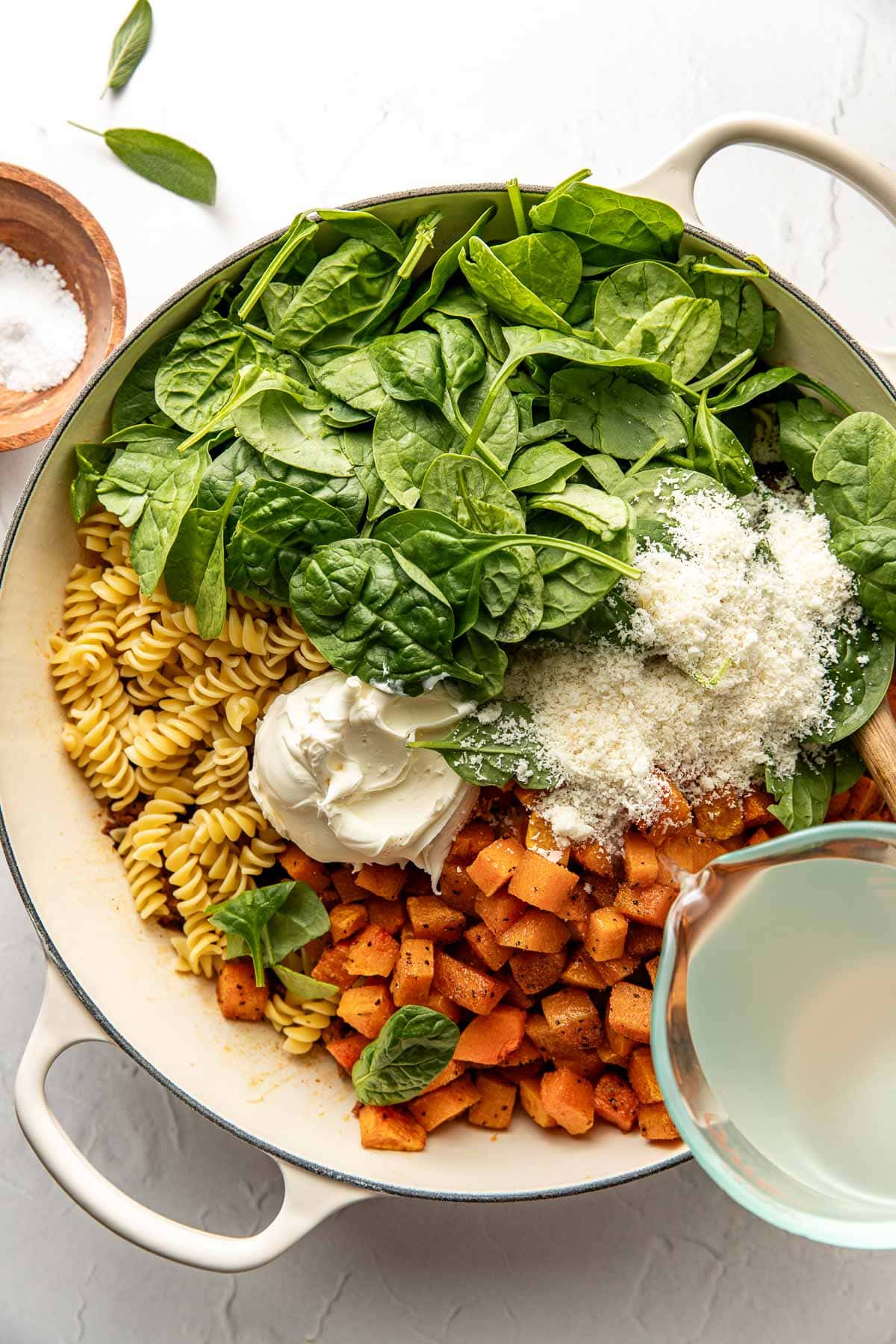 A large white pot filled with rotini pasta, fresh spinach, cubed roasted butternut squash, grated parmesan cheese, a dollop of cream cheese, and a small bowl of water being poured in.