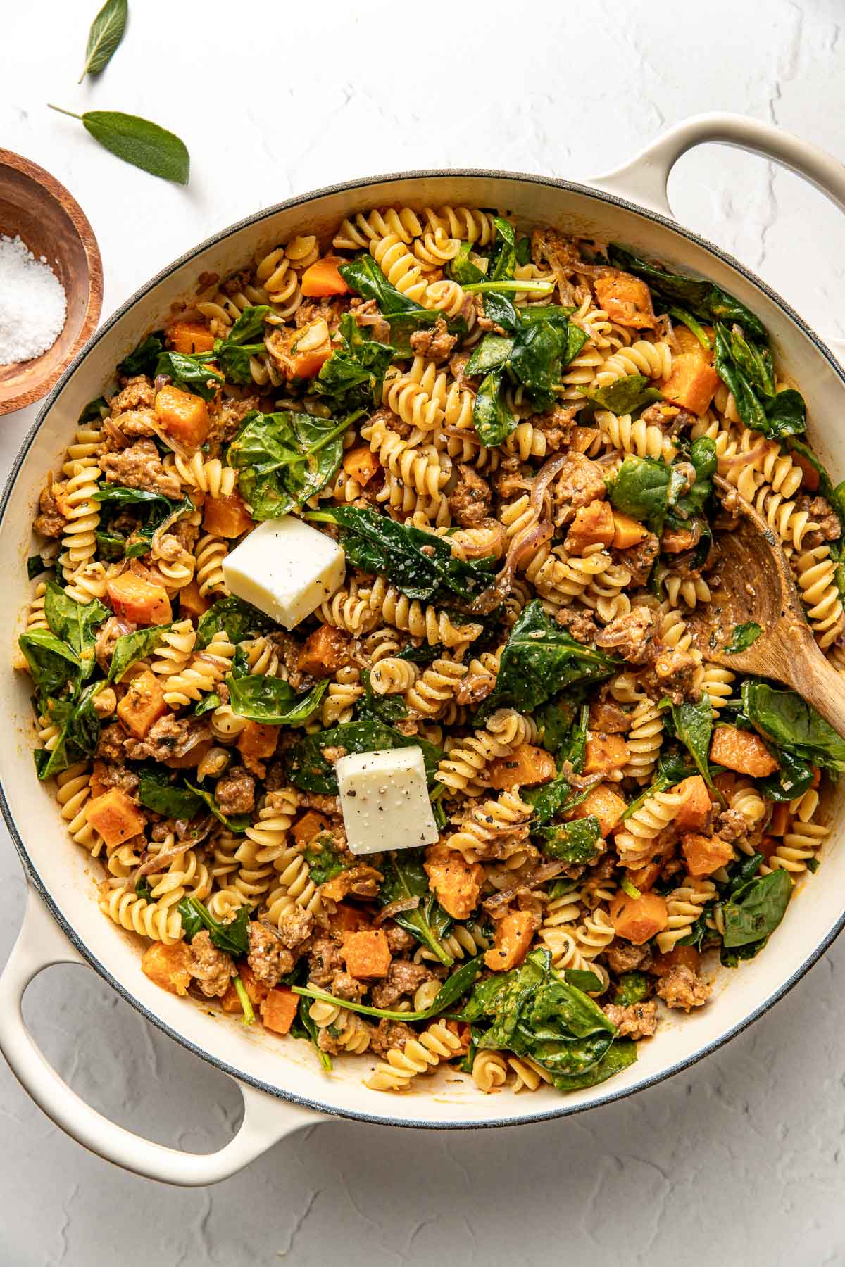 A large white skillet filled with rotini pasta, spinach, sausage, diced butternut squash, and two pats of butter, with a wooden spoon and a small bowl of salt on the side.