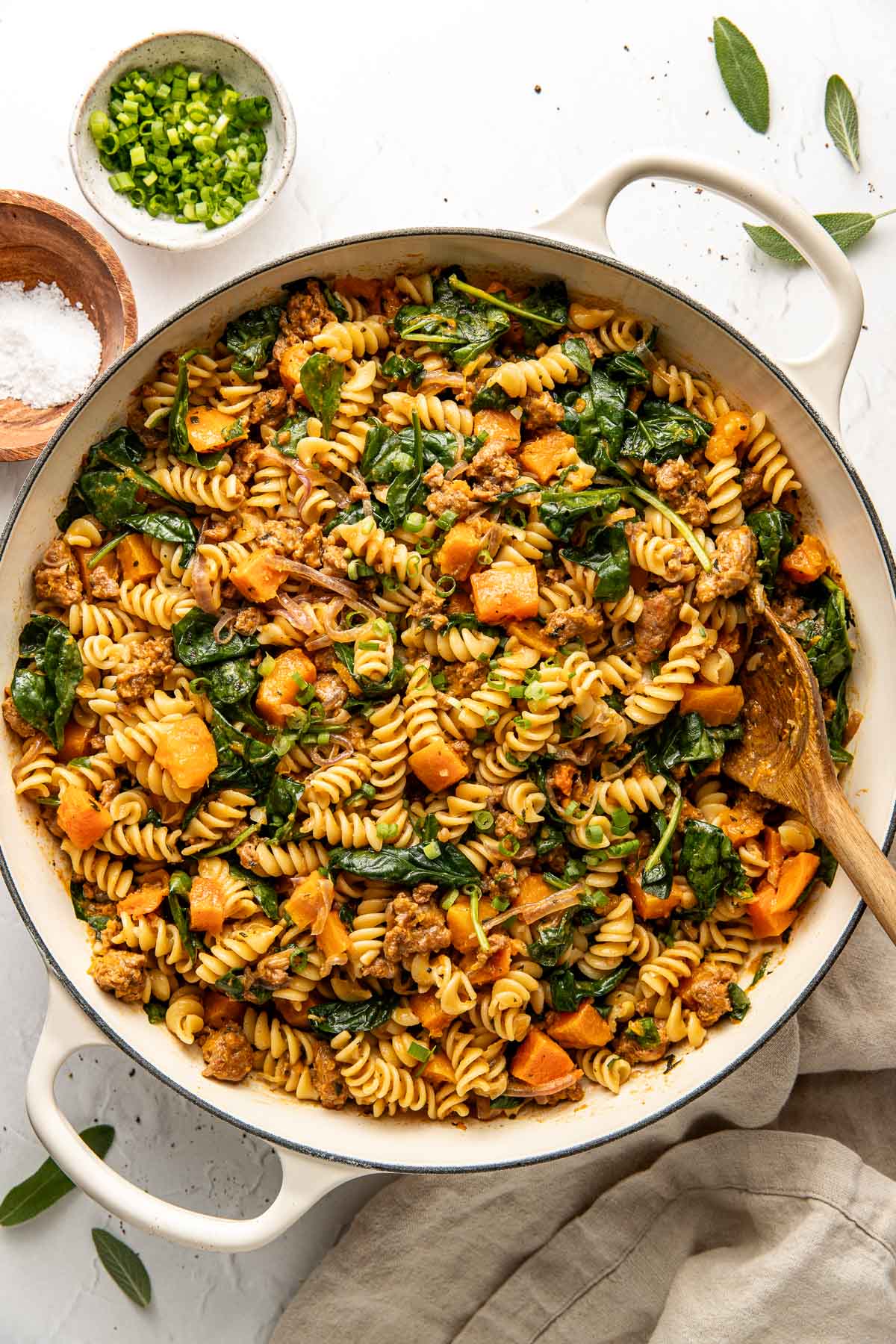 A large white pot filled with rotini pasta, cooked sausage, spinach, and chunks of butternut squash, mixed together with herbs. A wooden spoon rests in the dish, and chopped green onions are in a small bowl nearby.
