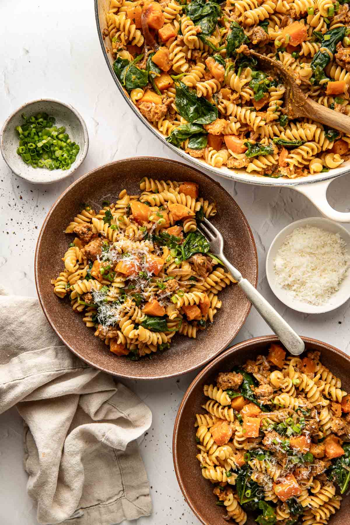 Two brown bowls filled with rotini pasta, sausage, spinach, and butternut squash sit beside a large skillet of the same dish. A bowl of grated cheese, chopped green onions, and a beige napkin are nearby.