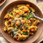 A bowl of rotini pasta mixed with sautéed greens, chunks of roasted butternut squash, sausage, and topped with grated cheese sits on a wooden board with a fork resting on the edge of the bowl.