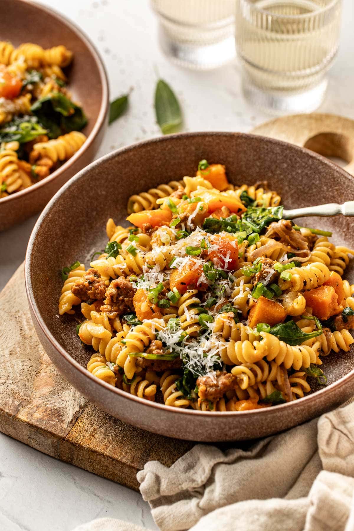 A bowl of rotini pasta mixed with ground meat, chopped kale, and roasted butternut squash, topped with grated cheese and herbs, sits on a wooden board next to a beige napkin. Another bowl and glasses of water are in the background.