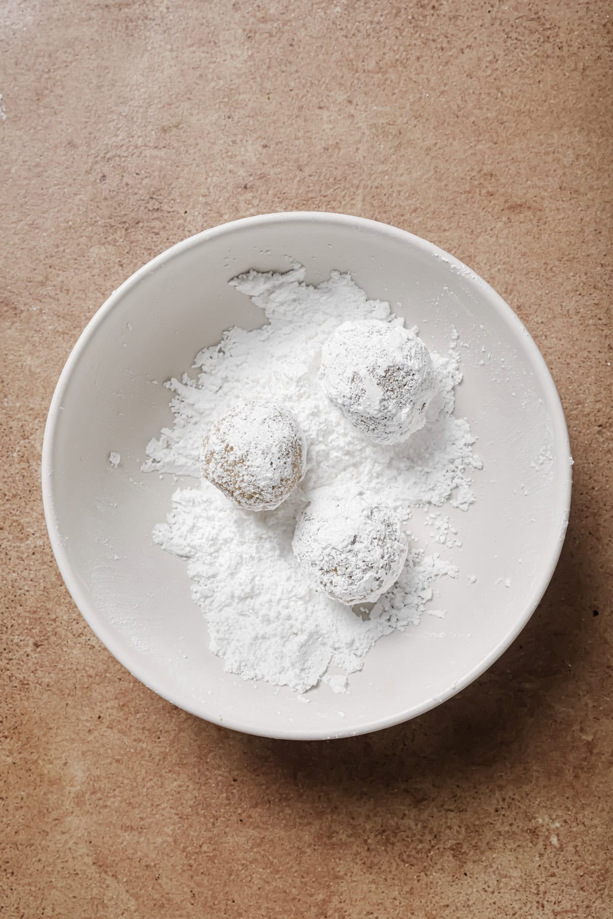 A white bowl with three round cookies partially coated in powdered sugar, sitting on a brown textured surface. Loose powdered sugar is scattered around the cookies in the bowl.
