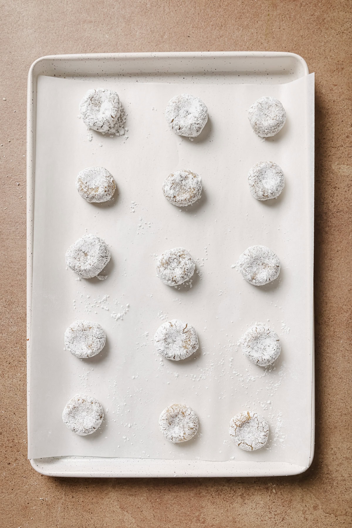 A baking tray lined with parchment paper holds fifteen evenly spaced, round cookies coated in powdered sugar, ready to be baked. The tray sits on a brown countertop.