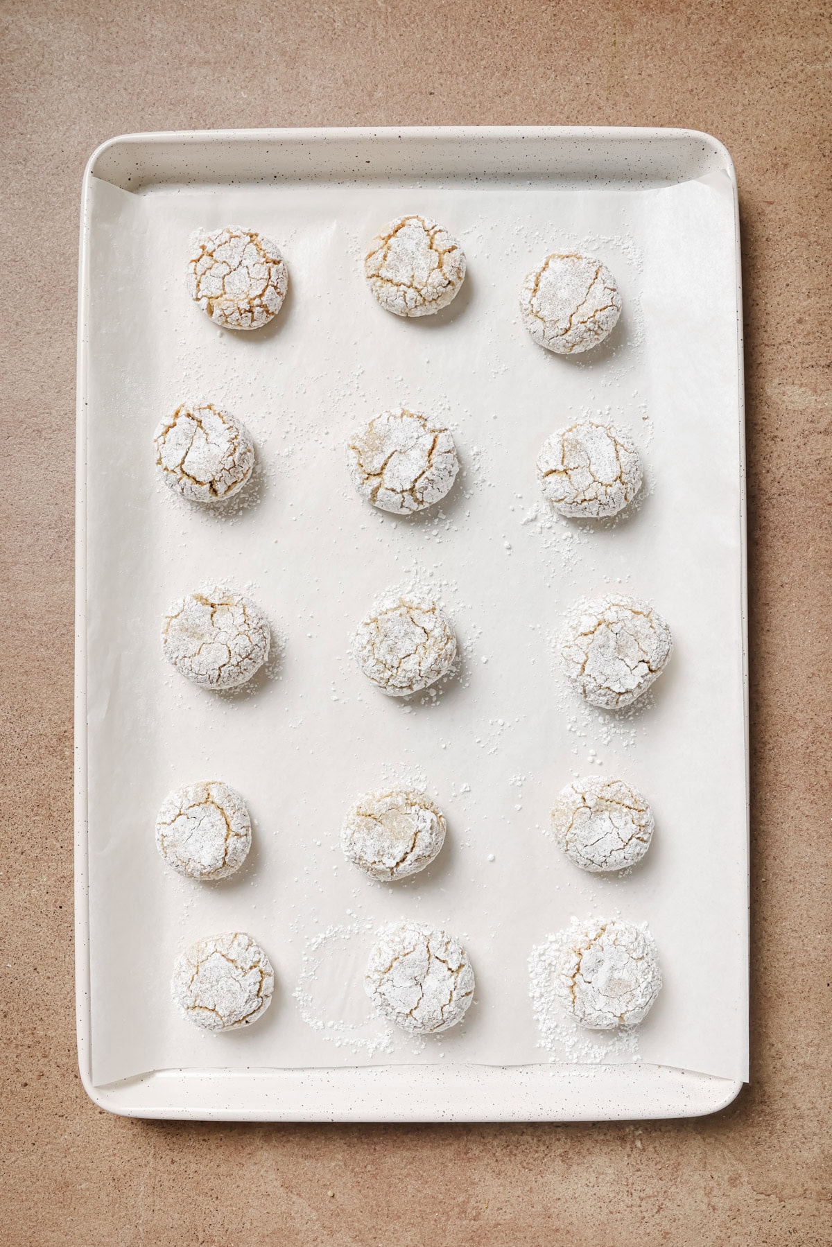 A baking tray lined with parchment paper holds fifteen round, cracked cookies dusted with powdered sugar, arranged in neat rows on a light brown surface.