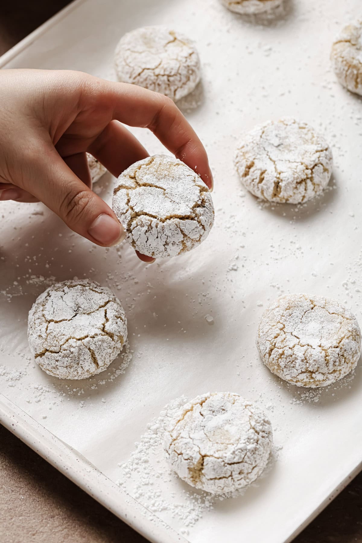 A hand picks up a powdered sugar-coated crinkle cookie from a parchment-lined baking sheet with several similar cookies, all lightly dusted with powdered sugar.