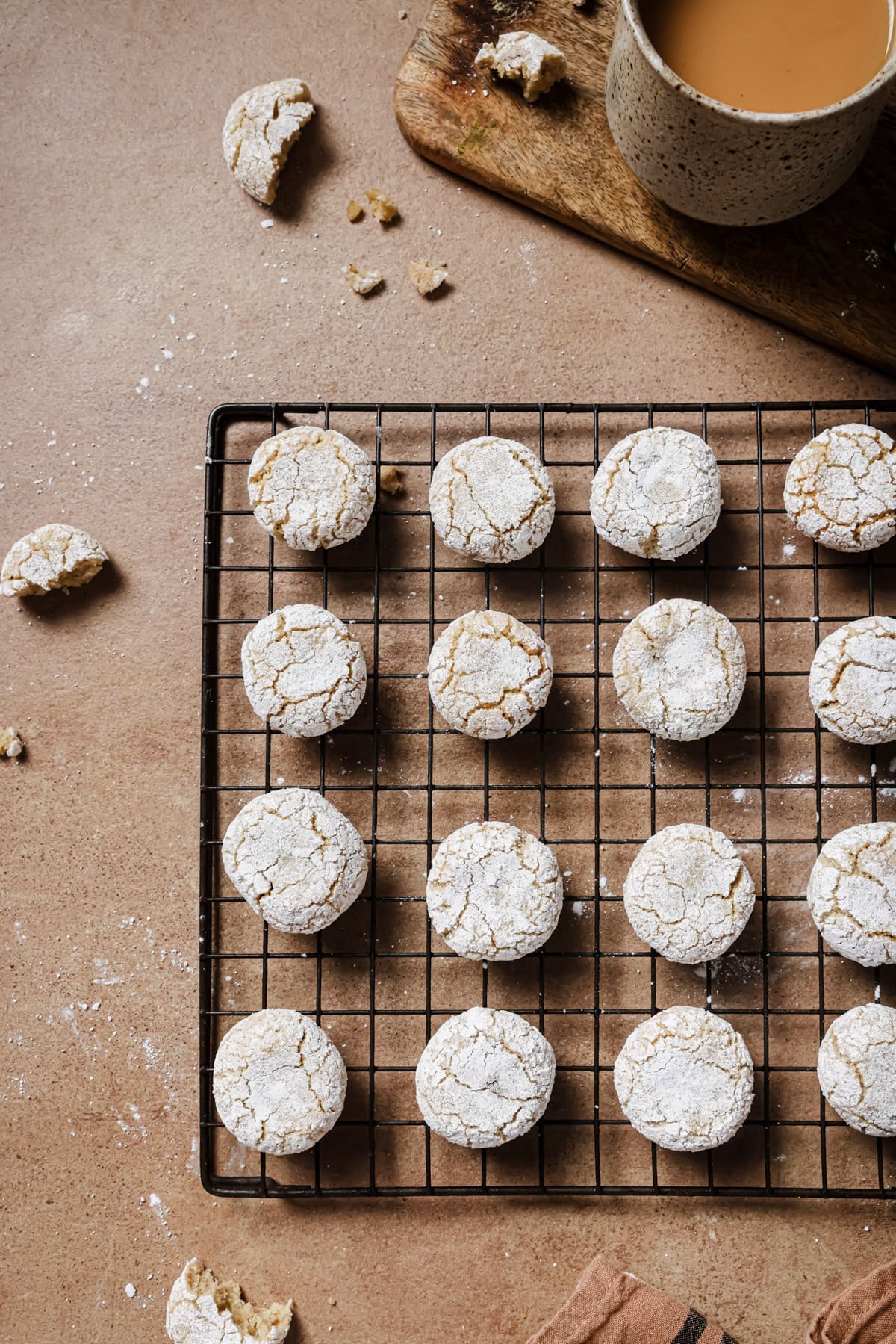 A cooling rack holds rows of powdered sugar-dusted cookies on a brown surface, with a cup of tea and cookie crumbs nearby.