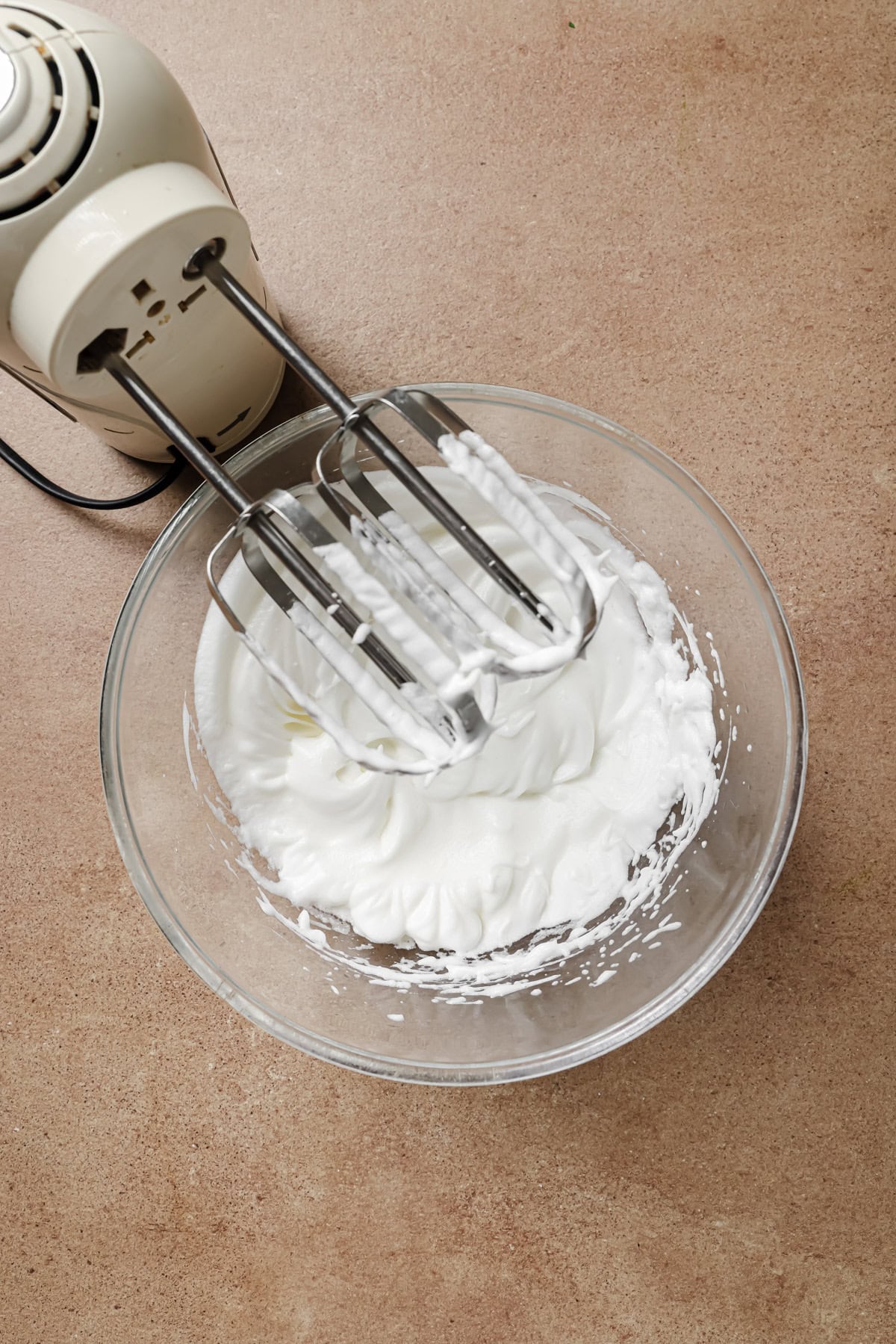 A hand mixer with metal beaters sits in a glass bowl containing whipped egg whites on a brown countertop.