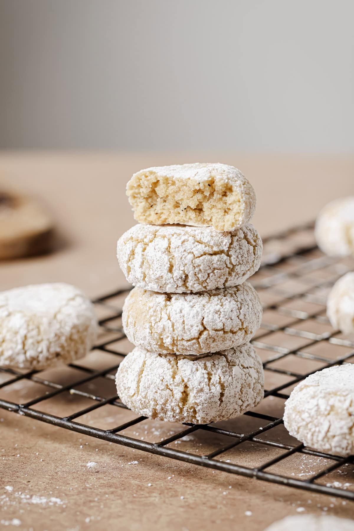A stack of four powdered sugar-coated cookies sits on a cooling rack, with the top cookie showing a bite taken out, revealing a soft, crumbly interior. Other cookies are scattered around on the rack and surface.