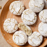 A round plate with powdered sugar-covered almond cookies, one broken in half to show a soft, crumbly interior. The cookies have a cracked surface and are arranged close together on a neutral background.