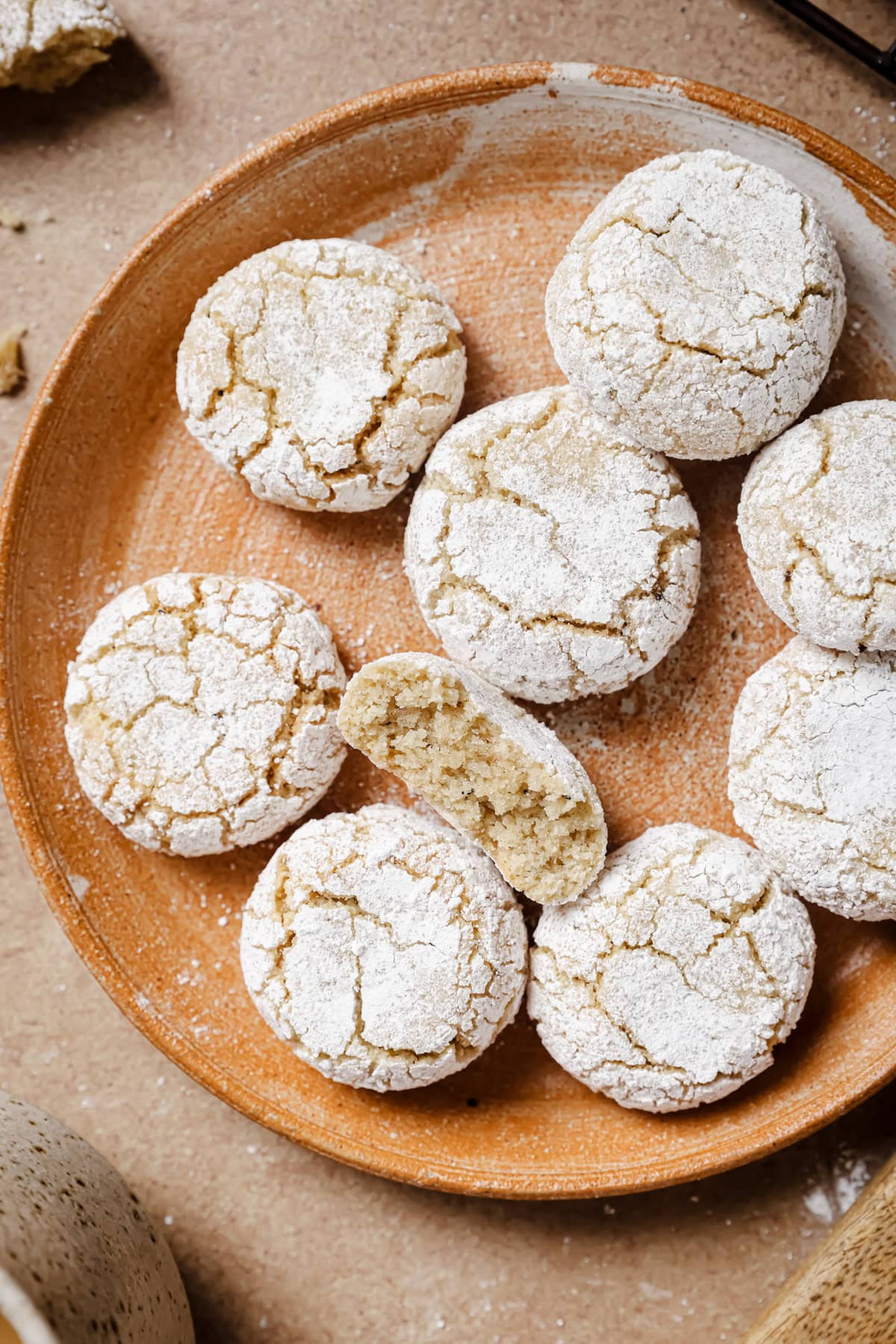 A round plate with powdered sugar-covered almond cookies, one broken in half to show a soft, crumbly interior. The cookies have a cracked surface and are arranged close together on a neutral background.