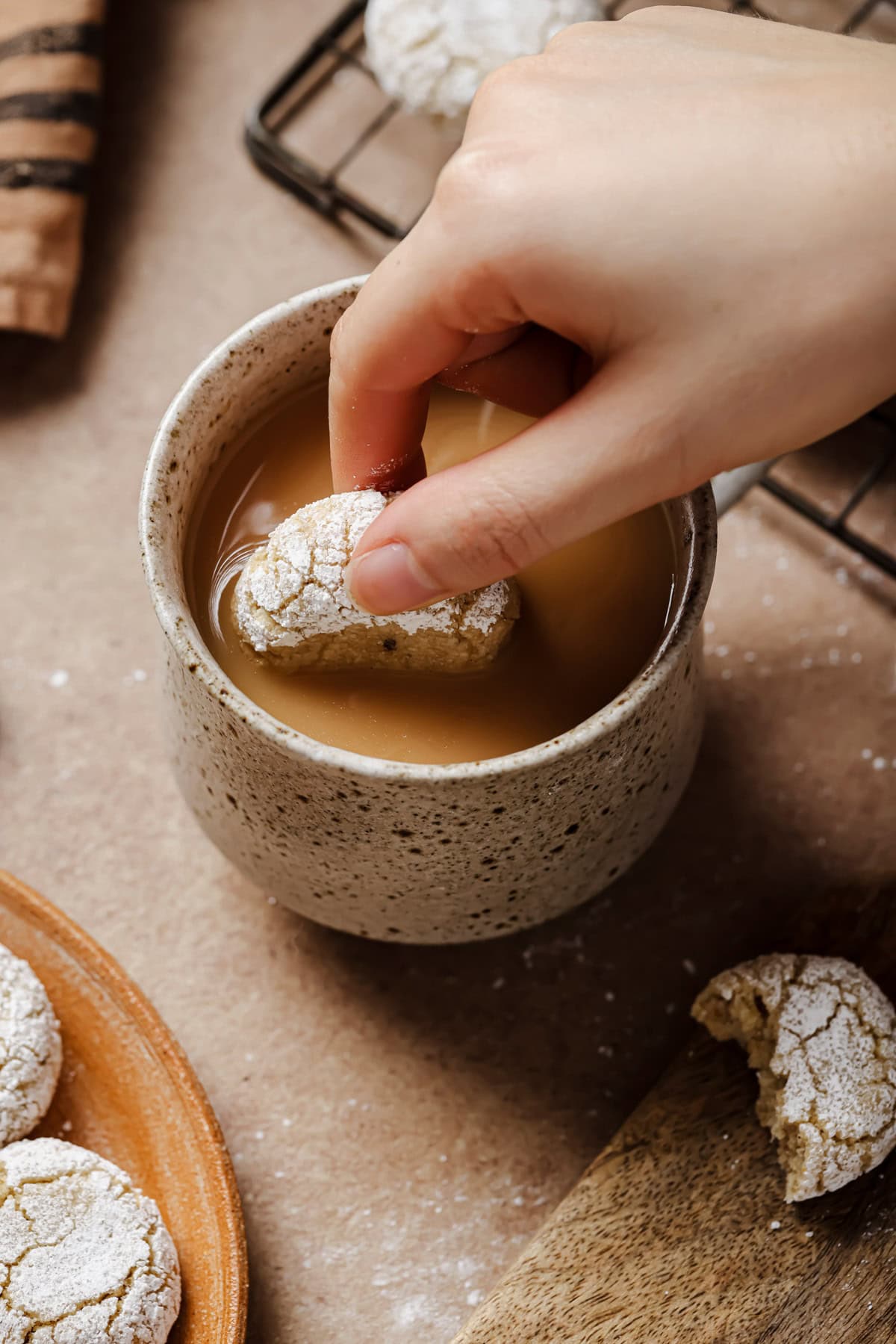A hand dips a powdered sugar cookie into a speckled ceramic mug filled with coffee or tea, with more cookies on a plate and wire rack nearby.