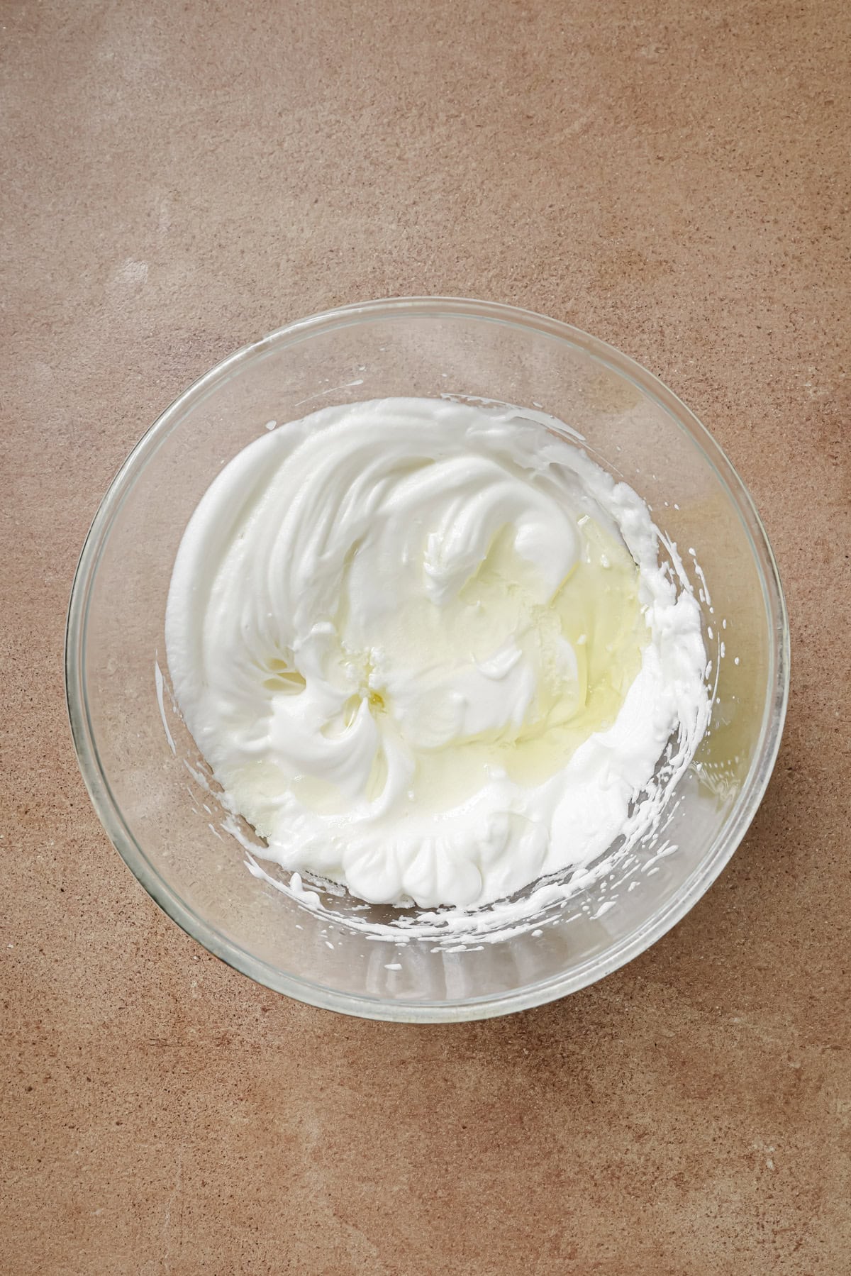 A glass bowl containing stiffly beaten egg whites with a glossy texture sits on a light brown countertop.