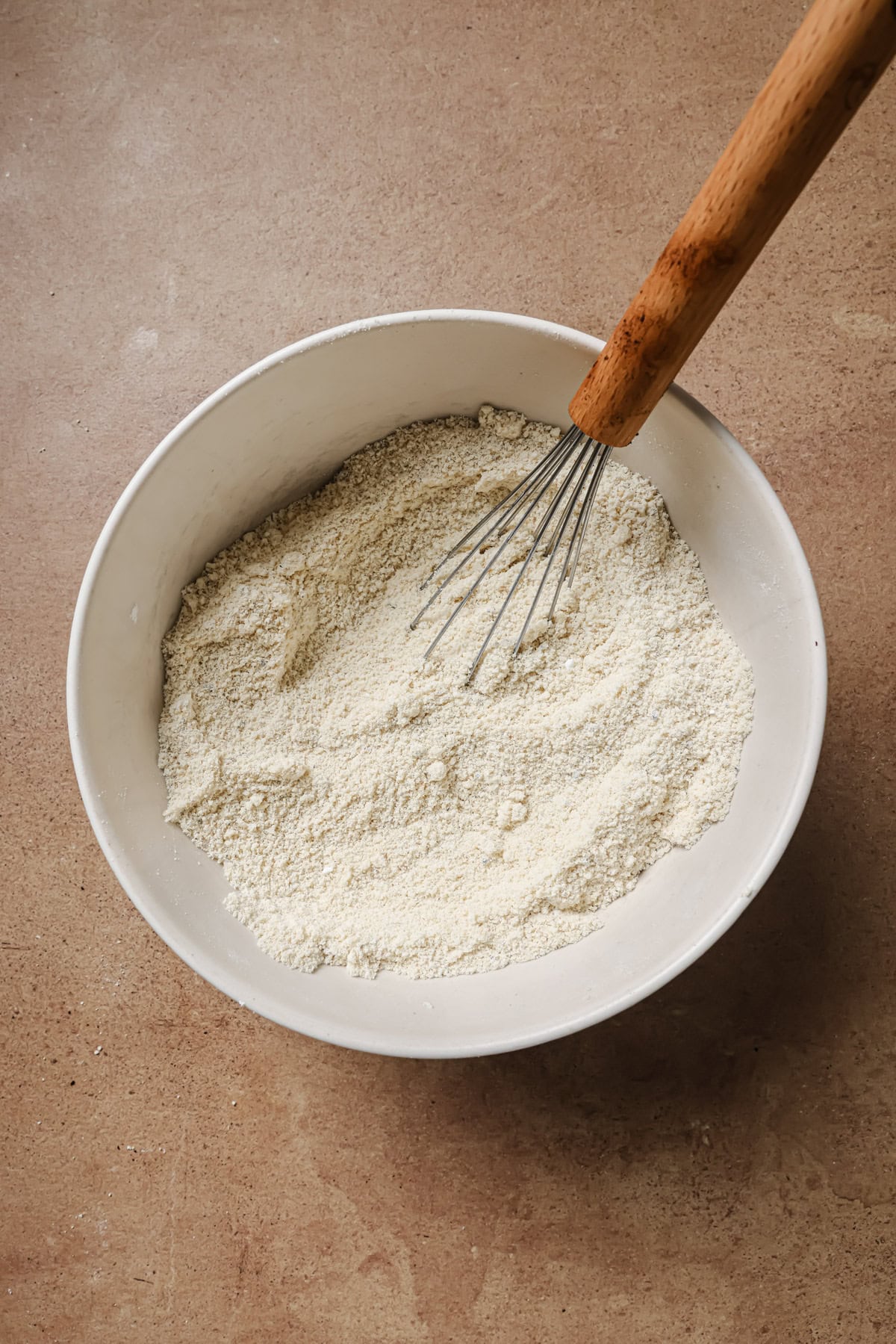 A white bowl filled with flour and a whisk resting inside, placed on a light brown countertop.