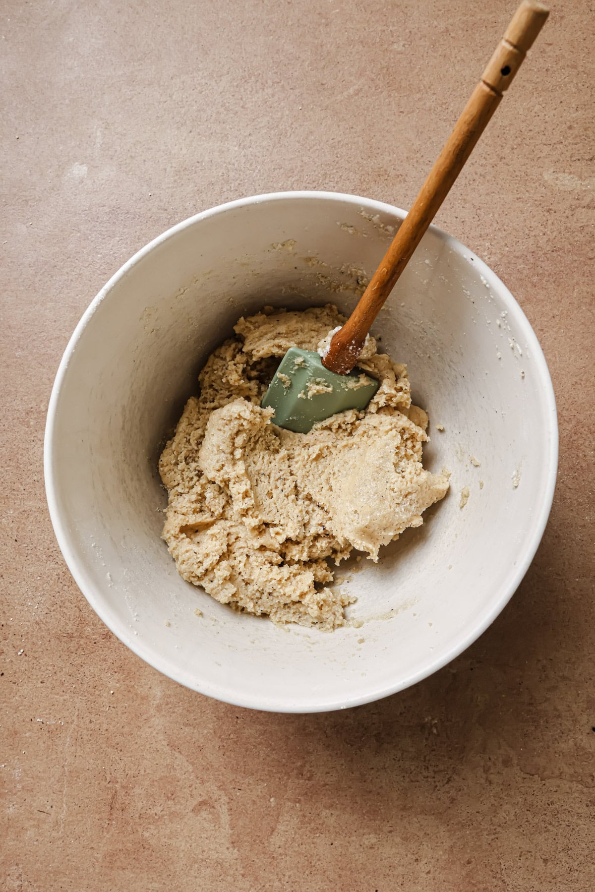 A white mixing bowl with thick cookie dough and a green spatula with a wooden handle, sitting on a brown countertop.