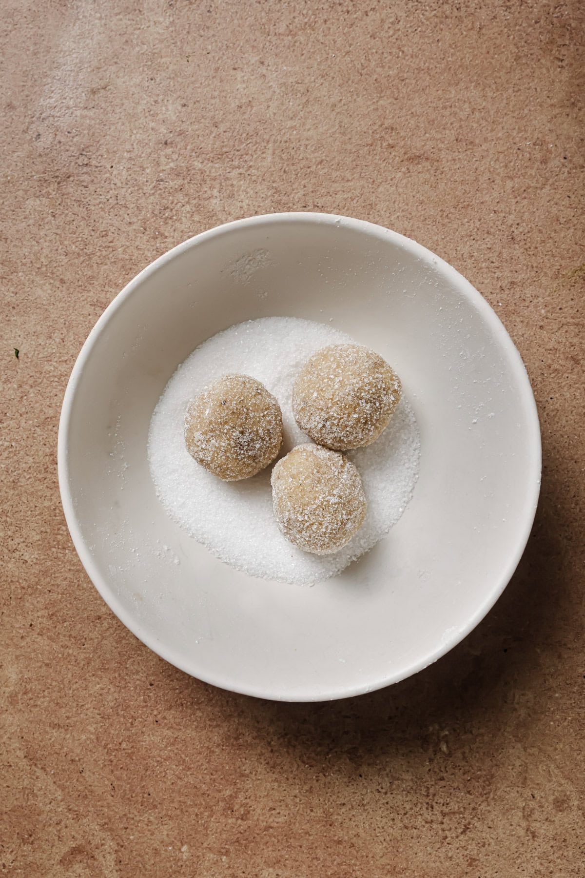 A white bowl containing three round, sugar-coated balls sits on a brown textured surface. The balls are partially resting in a layer of granulated sugar at the bottom of the bowl.