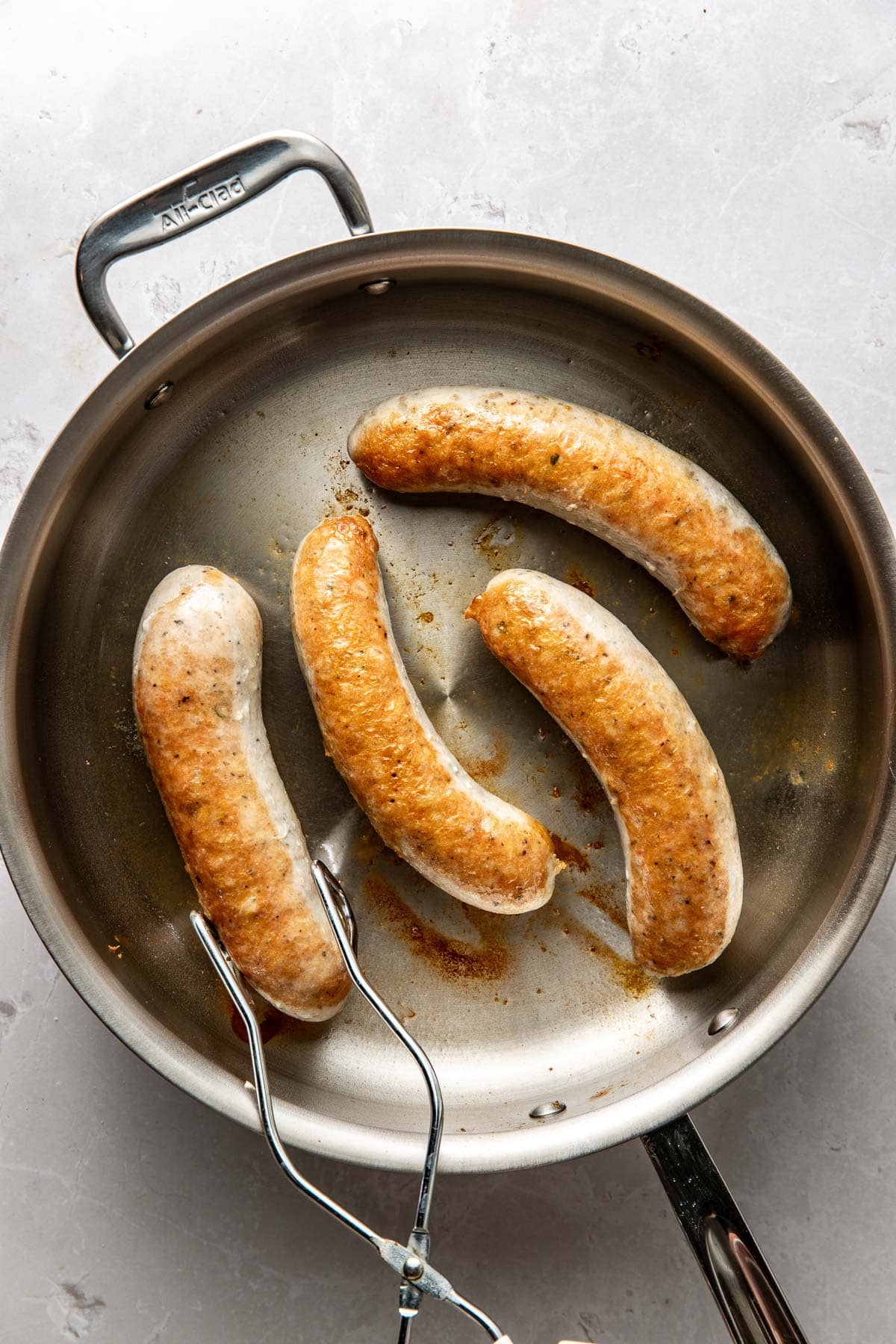 Four browned sausages cooking in a stainless steel skillet, with a pair of metal tongs holding one sausage, on a light-colored countertop.