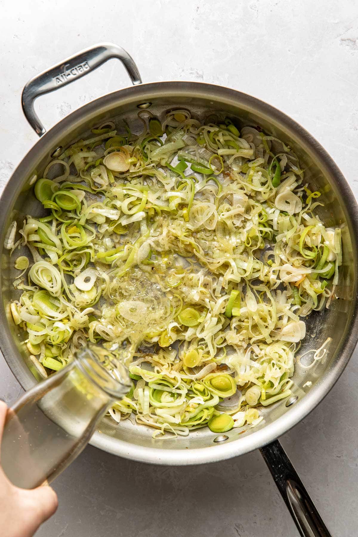 A hand pours liquid from a glass bottle into a pan of sliced leeks and onions cooking on a stovetop.
