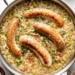 A skillet filled with creamy sauce, sautéed vegetables, and four browned sausages, garnished with chopped chives. Nearby are a bowl of chives, a bowl of salt, a glass of oil, and a beige cloth napkin.
