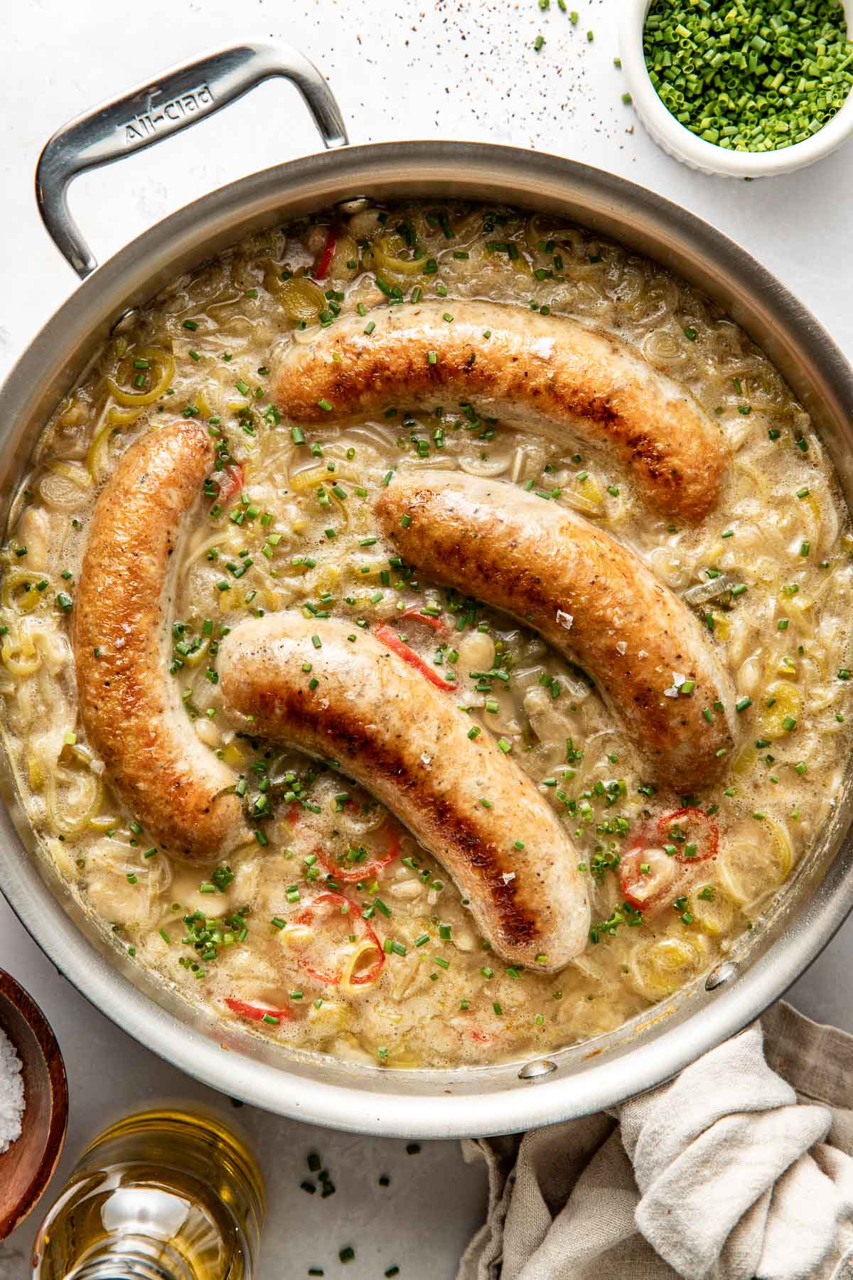 A skillet filled with creamy sauce, sautéed vegetables, and four browned sausages, garnished with chopped chives. Nearby are a bowl of chives, a bowl of salt, a glass of oil, and a beige cloth napkin.