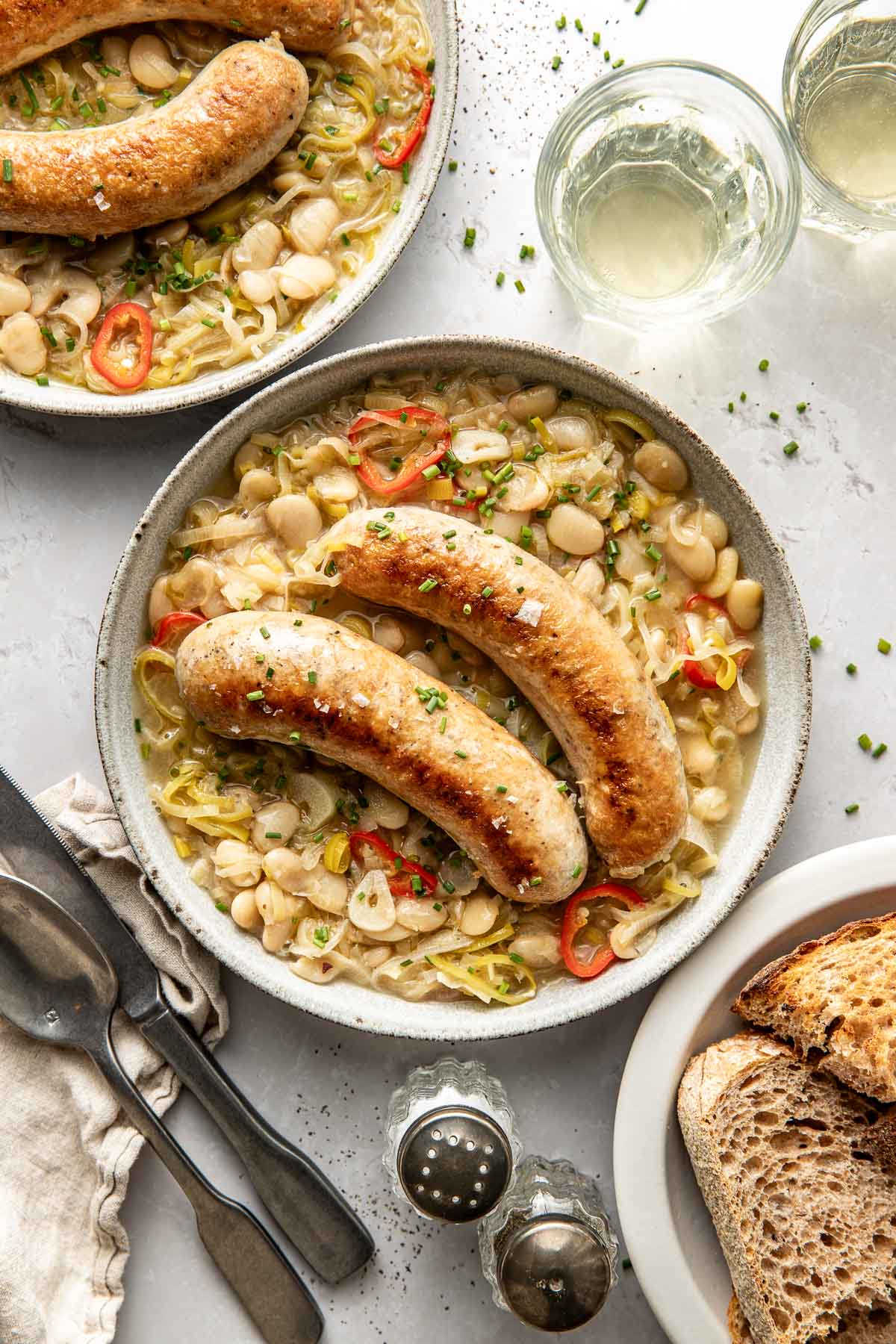 A bowl with two browned sausages atop a creamy bean and leek mixture, garnished with sliced red chili and chives. Slices of rustic bread and glasses of white wine are beside the plate.