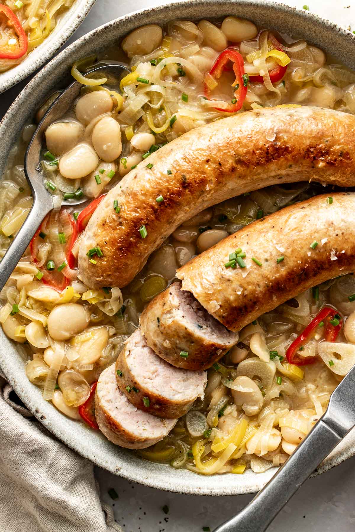 A bowl of white beans and vegetables topped with two grilled sausages, one sliced, garnished with chopped herbs and red pepper rings. A fork rests in the bowl next to the food.