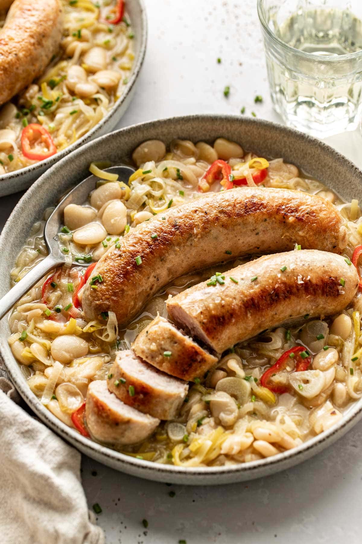 A bowl of creamy white bean stew with sliced and whole sausages on top, garnished with herbs and sliced red chili peppers. A glass of water and a napkin are next to the bowl.