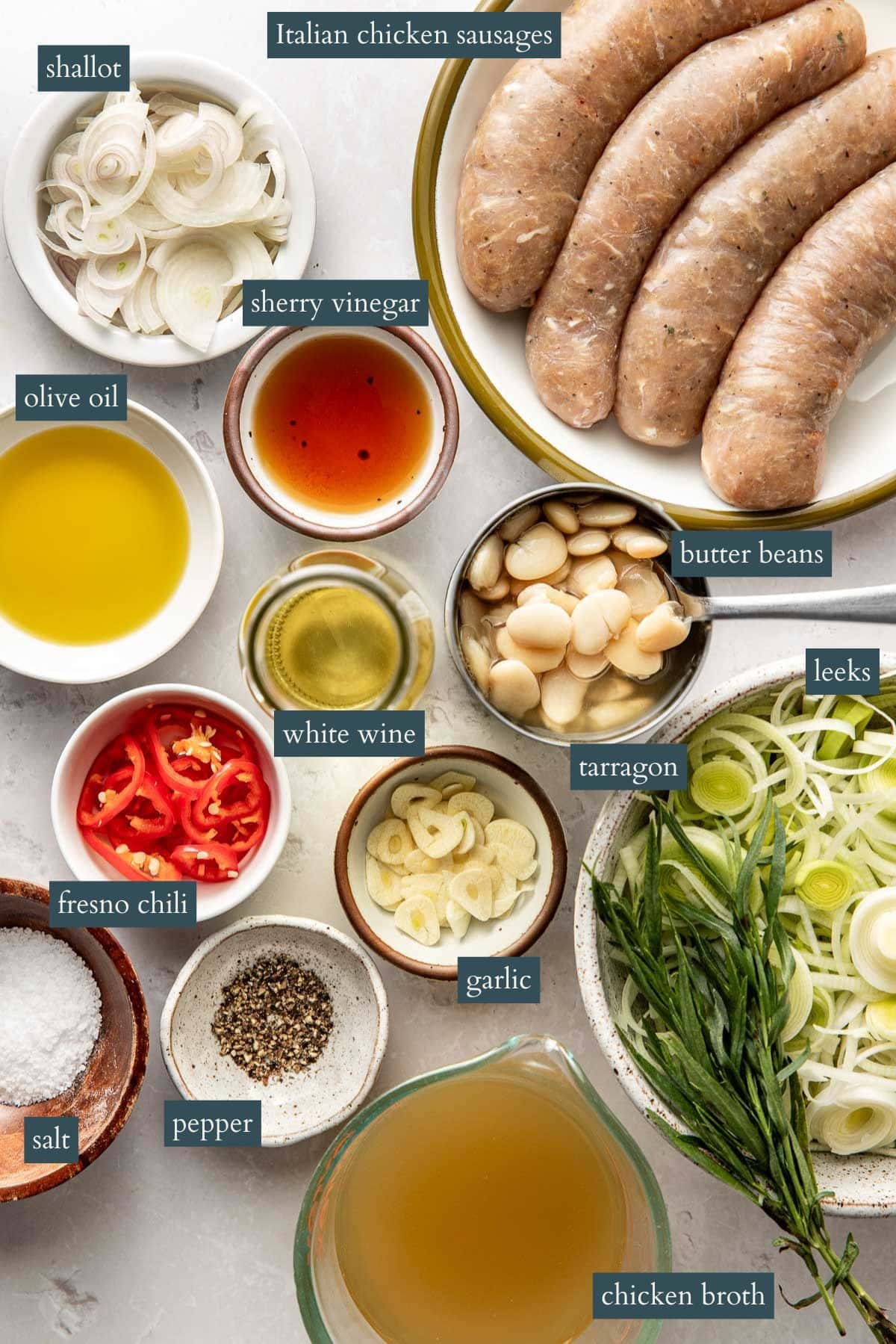 A top view of ingredients in bowls and plates, including Italian chicken sausages, sliced shallots, olive oil, sherry vinegar, white wine, butter beans, leeks, tarragon, Fresno chili, garlic, salt, pepper, and chicken broth. Labels identify each item.