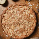 A round almond tart topped with sliced almonds sits on a wooden surface, with a small bowl of extra almonds, a fork, and a halved pear nearby. Crumbs are scattered around the tart.