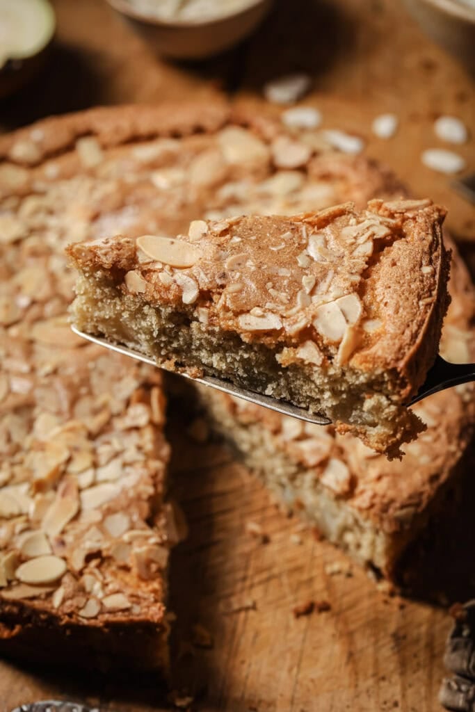 A close-up of a slice of almond cake topped with sliced almonds being lifted from the whole cake on a wooden surface.