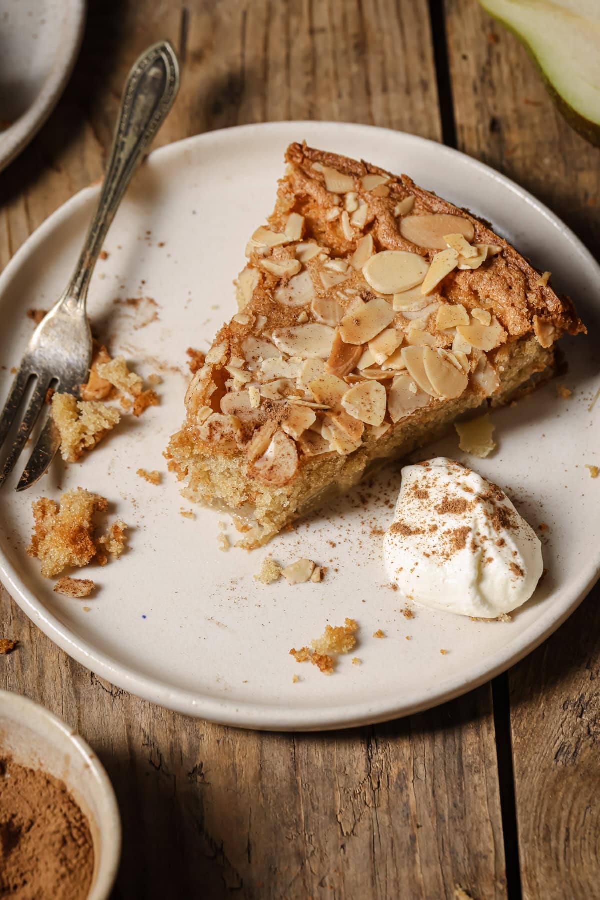 A slice of almond-topped cake sits on a white plate with a dollop of whipped cream and a dusting of cinnamon. A fork and crumbs are on the rustic wooden table beside the plate.