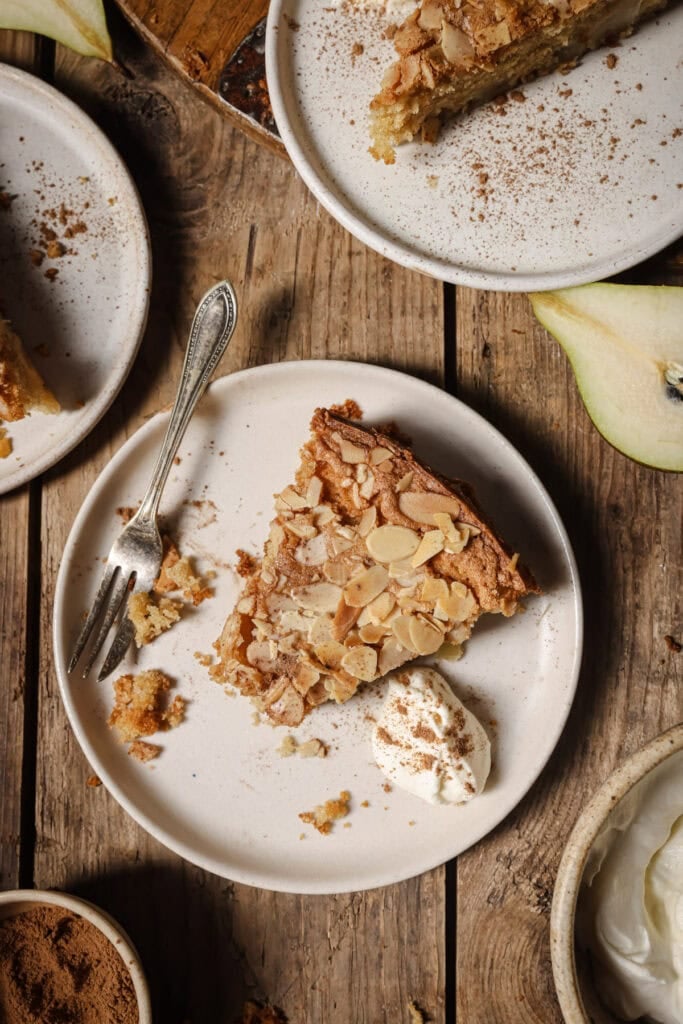 A slice of almond-topped cake sits on a plate with a dollop of cream and a fork. Surrounding it are more plates with cake, a pear slice, and a wooden table dusted with cinnamon.