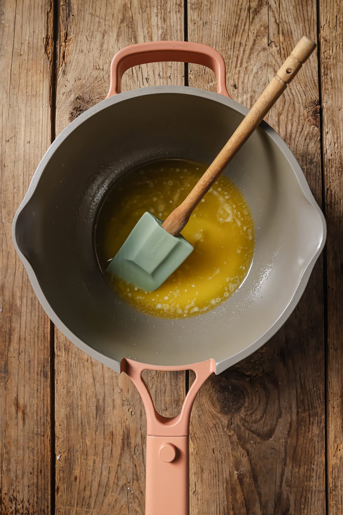 A light pink pot with a melted yellow substance and a green spatula sits on a rustic wooden surface.