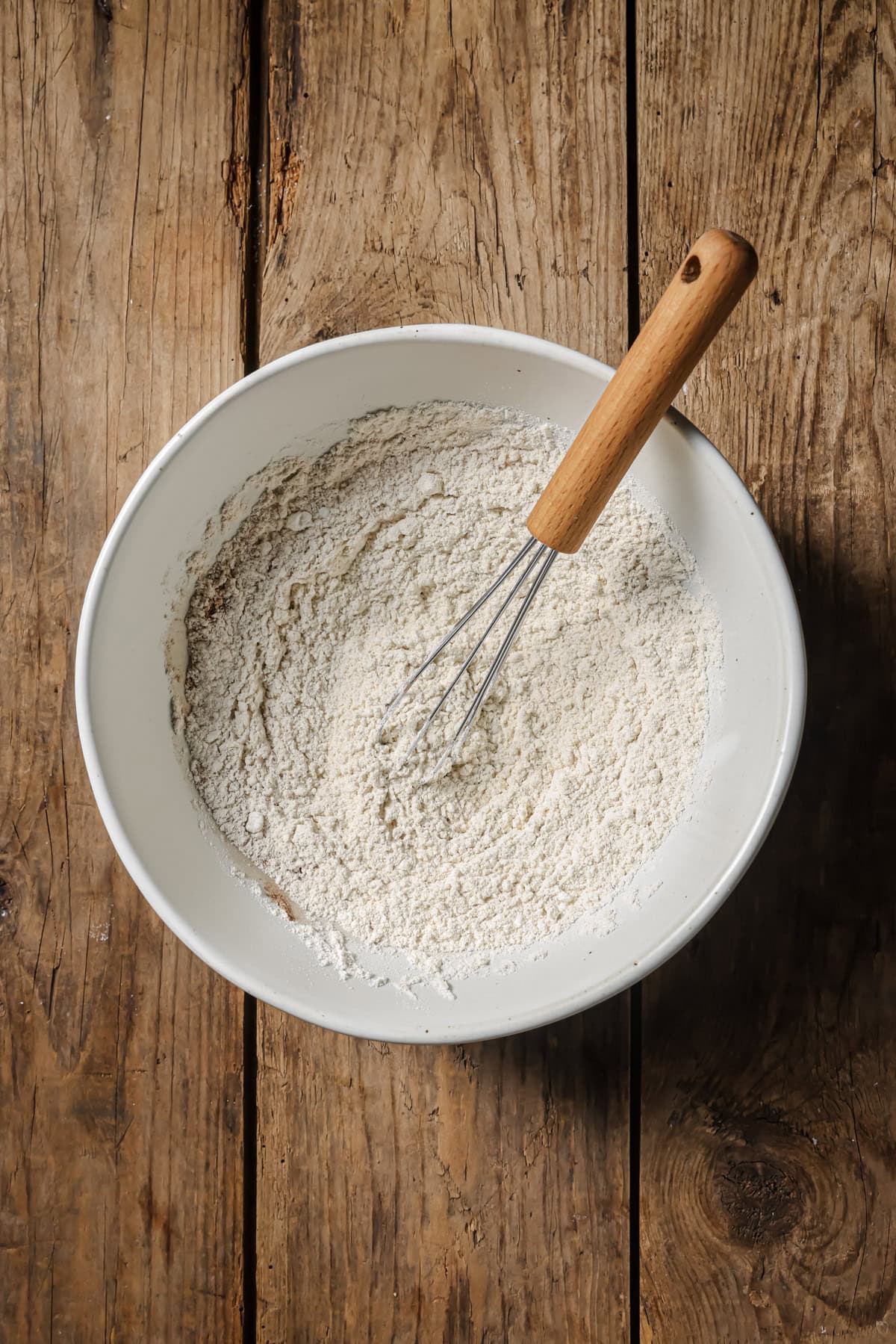 A white ceramic bowl filled with flour and a metal whisk with a wooden handle sits on a rustic wooden surface.
